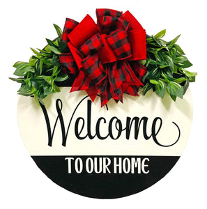 Painted 18 inch wood round door hanger. black background with welcome to our home text on a large white stripe. red and black plaid bow with greenery.