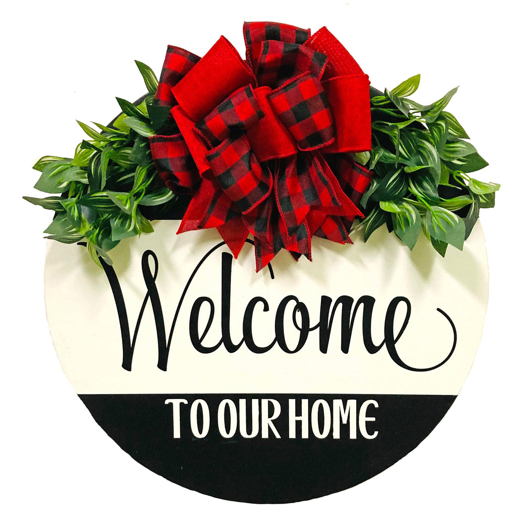 Painted 18 inch wood round door hanger. black background with welcome to our home text on a large white stripe. red and black plaid bow with greenery.
