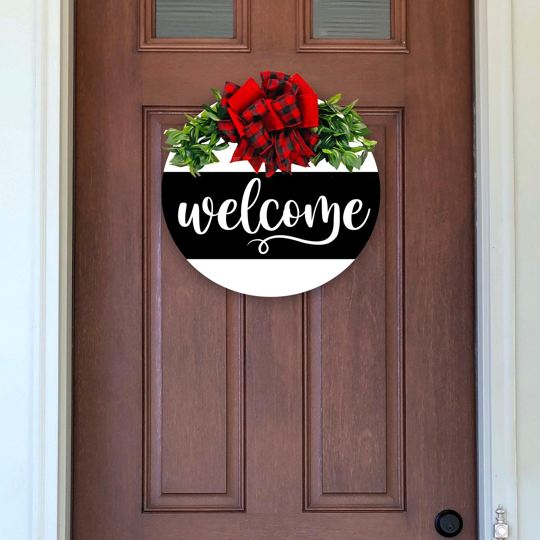 Painted 18 inch wood round door hanger. white background with welcome text on a large black stripe. black and white plaid bow with greenery.