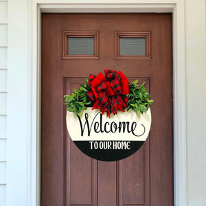 Painted 18 inch wood round door hanger. black background with welcome to our home text on a large white stripe. red and black plaid bow with greenery