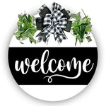 Painted 18 inch wood round door hanger.  white background with welcome text on a large black stripe.  black and white plaid bow with greenery.